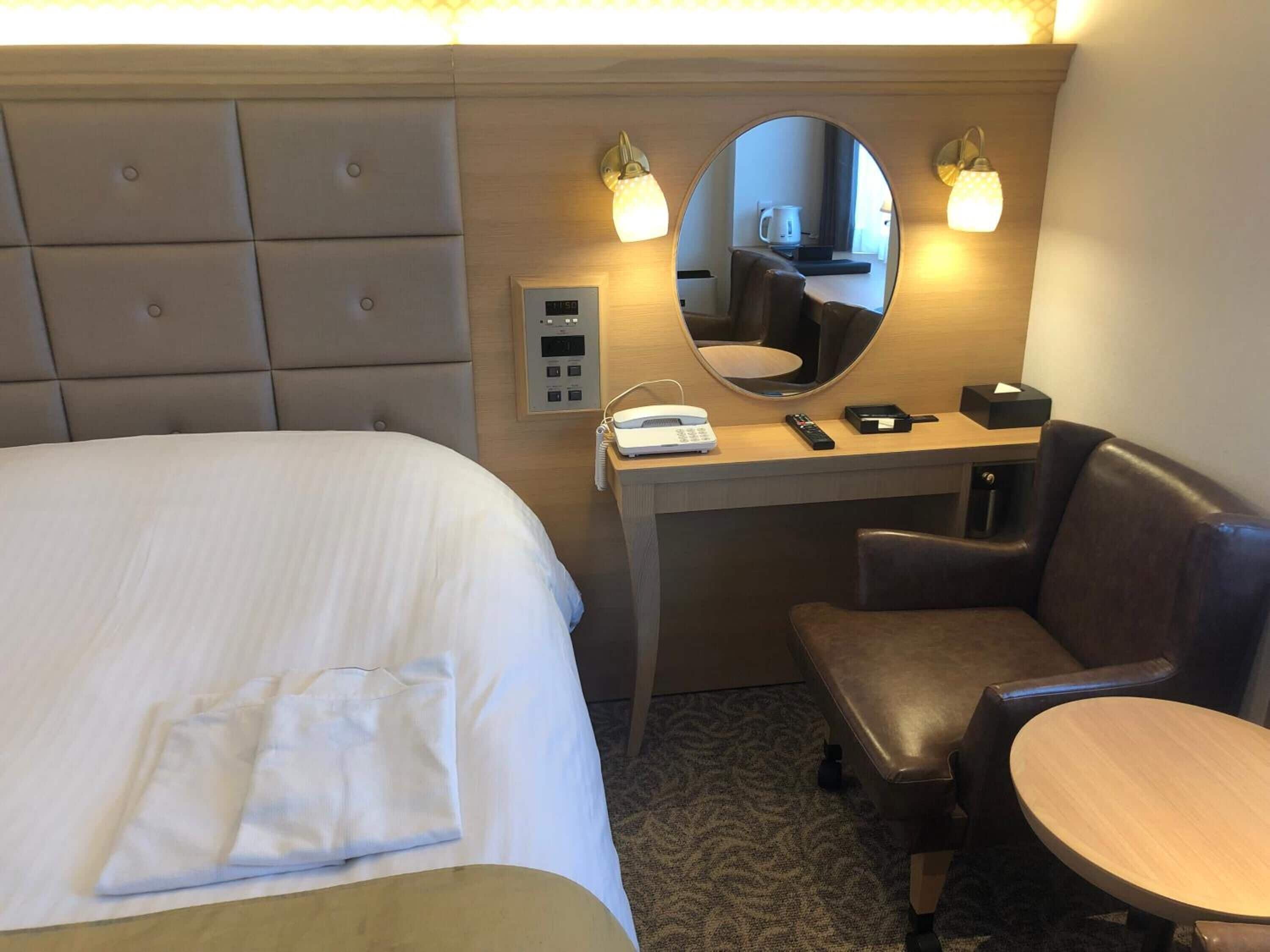 A hotel room with a bed, a desk with a mirror, a chair, and a small table.
