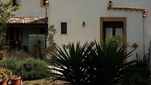 A building with a flat roof, a small window, and a door. There are potted plants and a large plant in the foreground. - B&B Le Macine 1868 (Paceco)