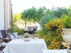 An outdoor patio with a table, chairs, and a bench. There are potted plants and yellow flowers in the background.