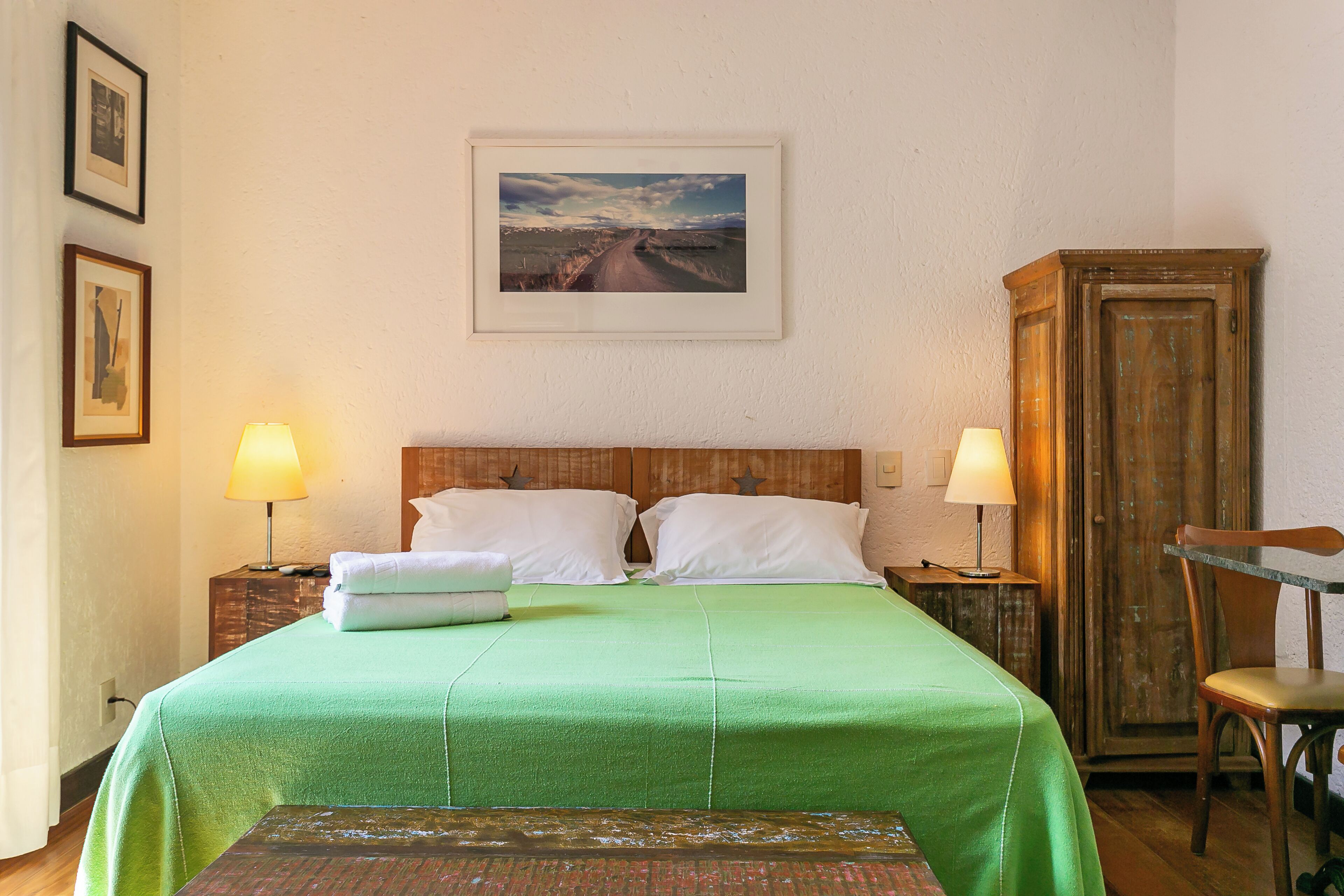 A neatly made bed with a green bedspread, two bedside lamps, a framed picture on the wall, and a wooden cabinet.