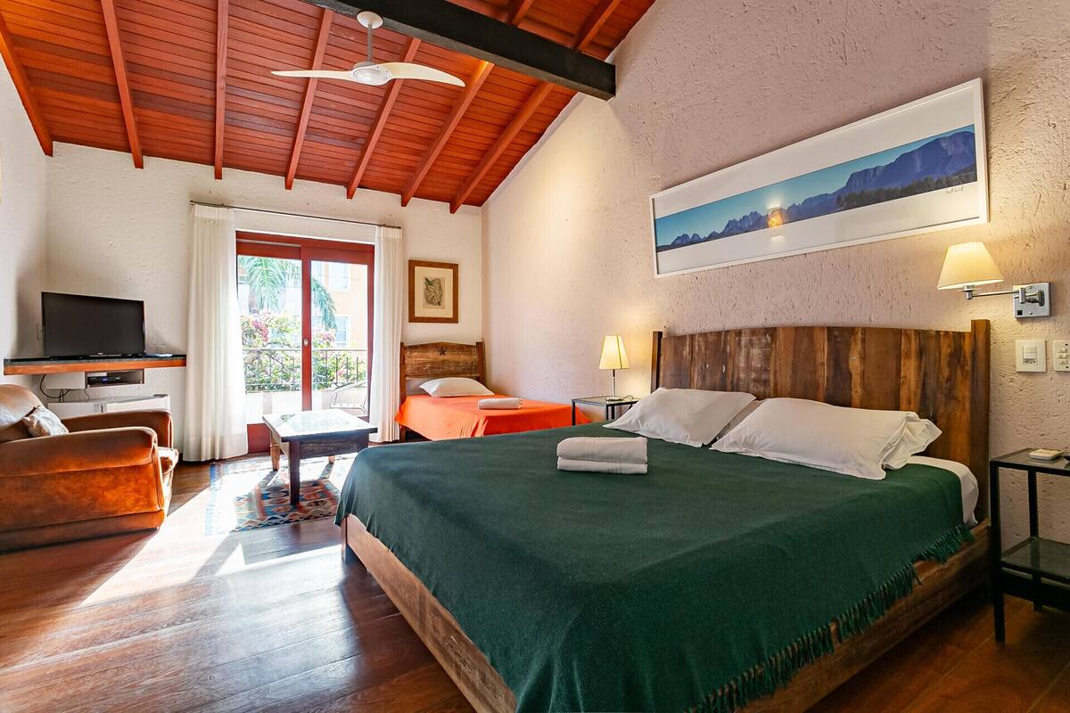 Ein Hotelzimmer mit Holzboden, einem Bett mit grüner Bettdecke, einem Schreibtisch mit Fernseher, einem Sessel und einem Fenster mit Bergblick.