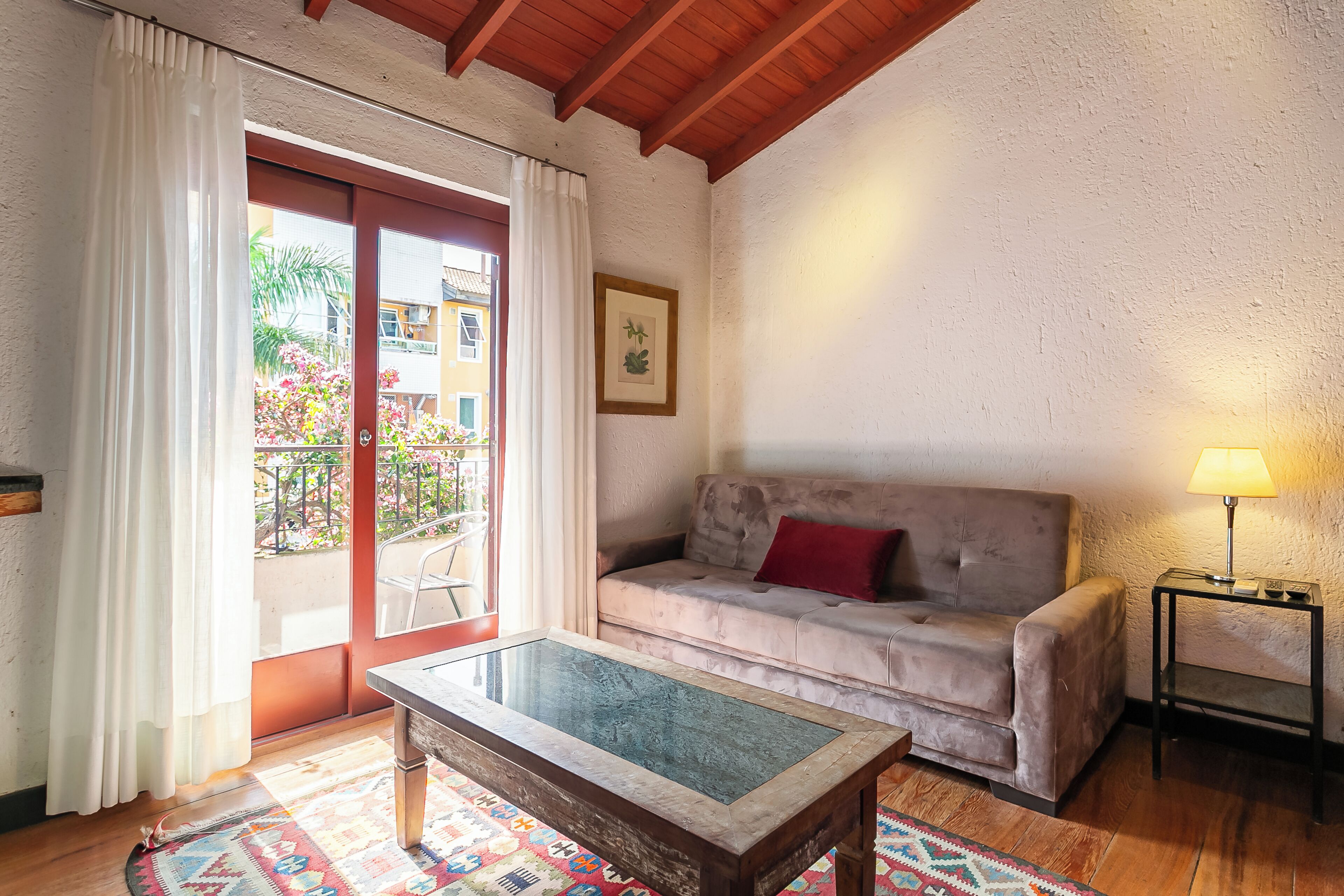 Salón con una mesa de centro de vidrio, un sofá con un cojín rojo, una lámpara sobre una mesita auxiliar y vistas a un espacio exterior con plantas a través de una puerta corrediza de vidrio.