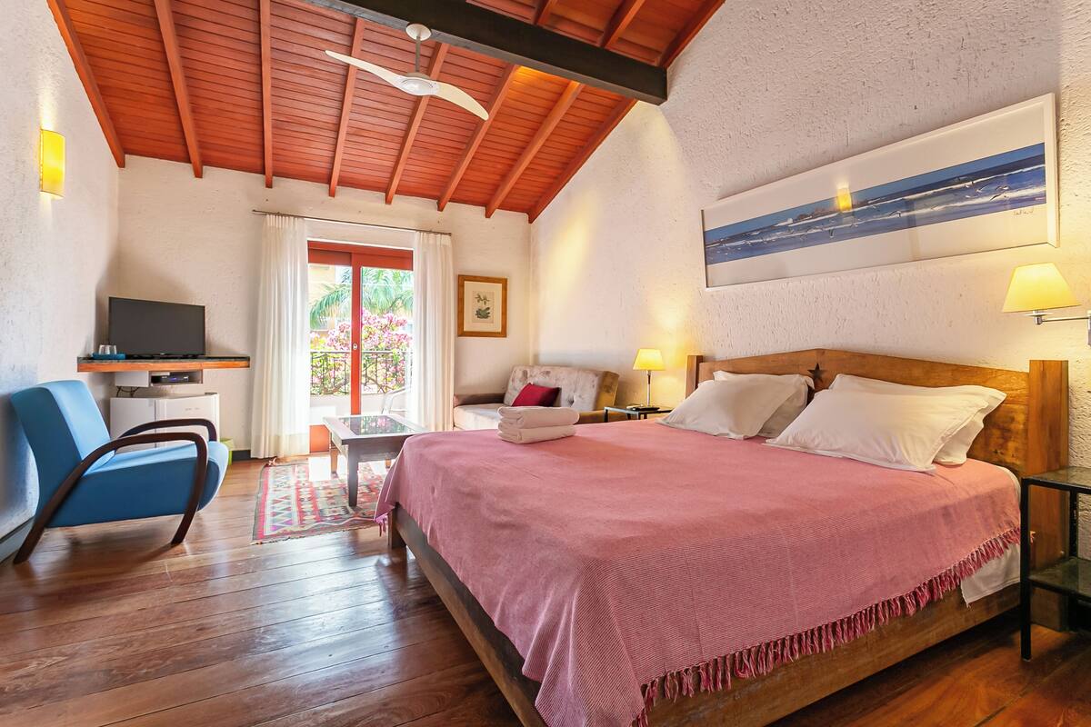 Ein Schlafzimmer mit Holzboden, einem Bett mit rosa Bettdecke, einem Fernseher auf einem Beistelltisch, einem blauen Sessel und einem Fenster mit Vorhängen.