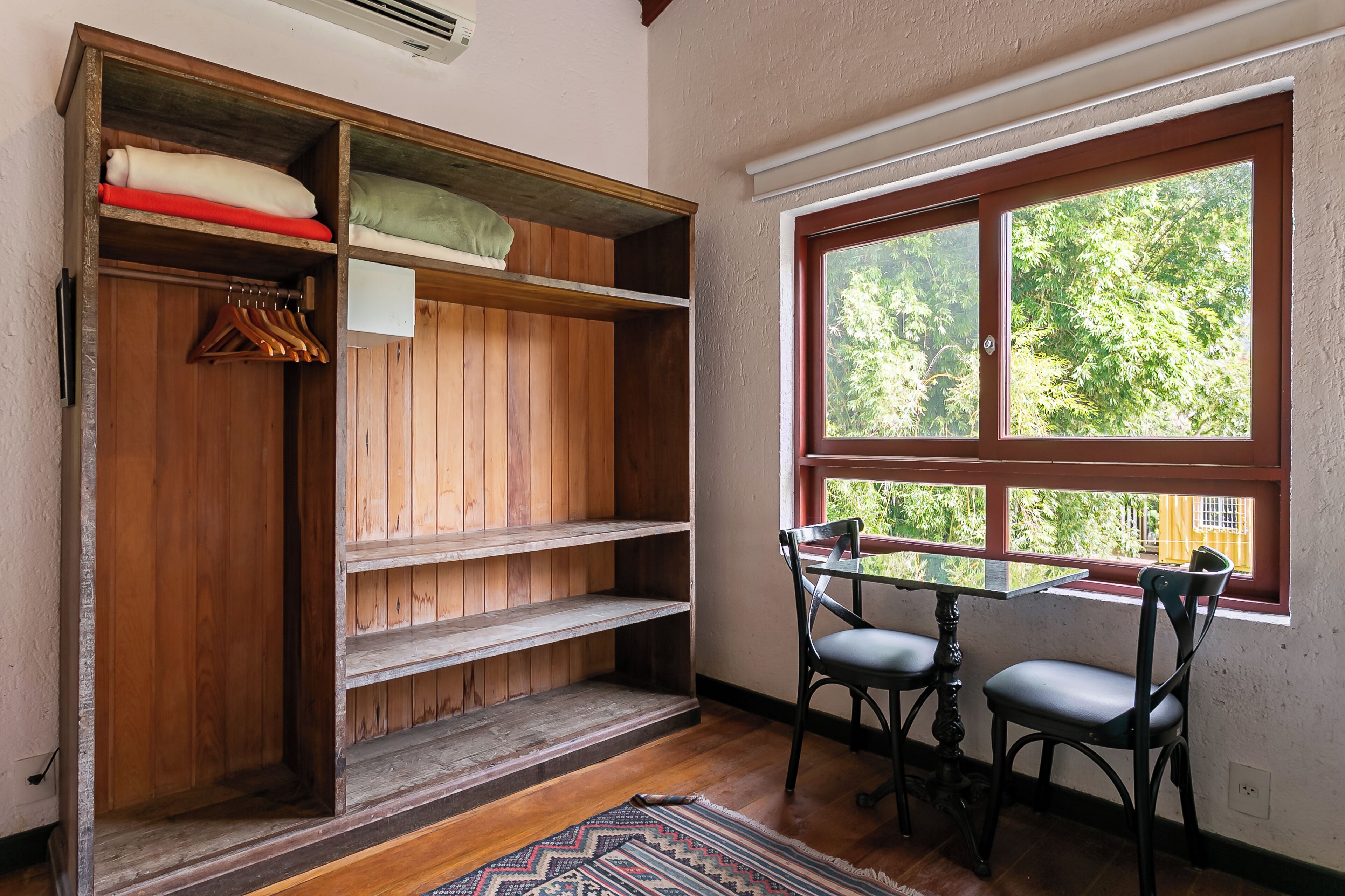 Ein Zimmer mit einem Holzschrank, einem Tisch mit zwei Stühlen und einem Fenster mit Blick auf Bäume.