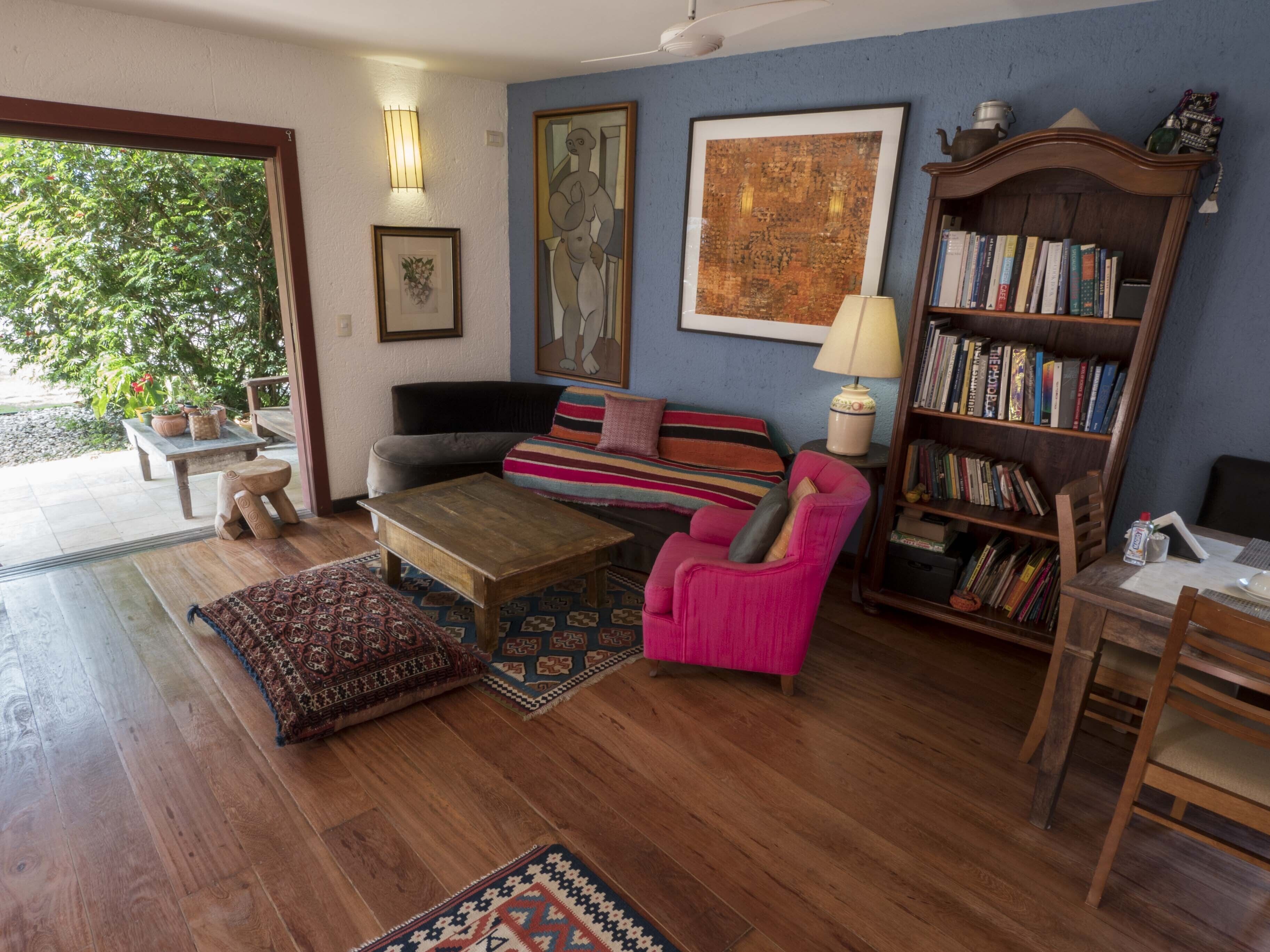 A cozy living room with a sofa, a coffee table, a pink armchair, a bookshelf, and a large window overlooking greenery.