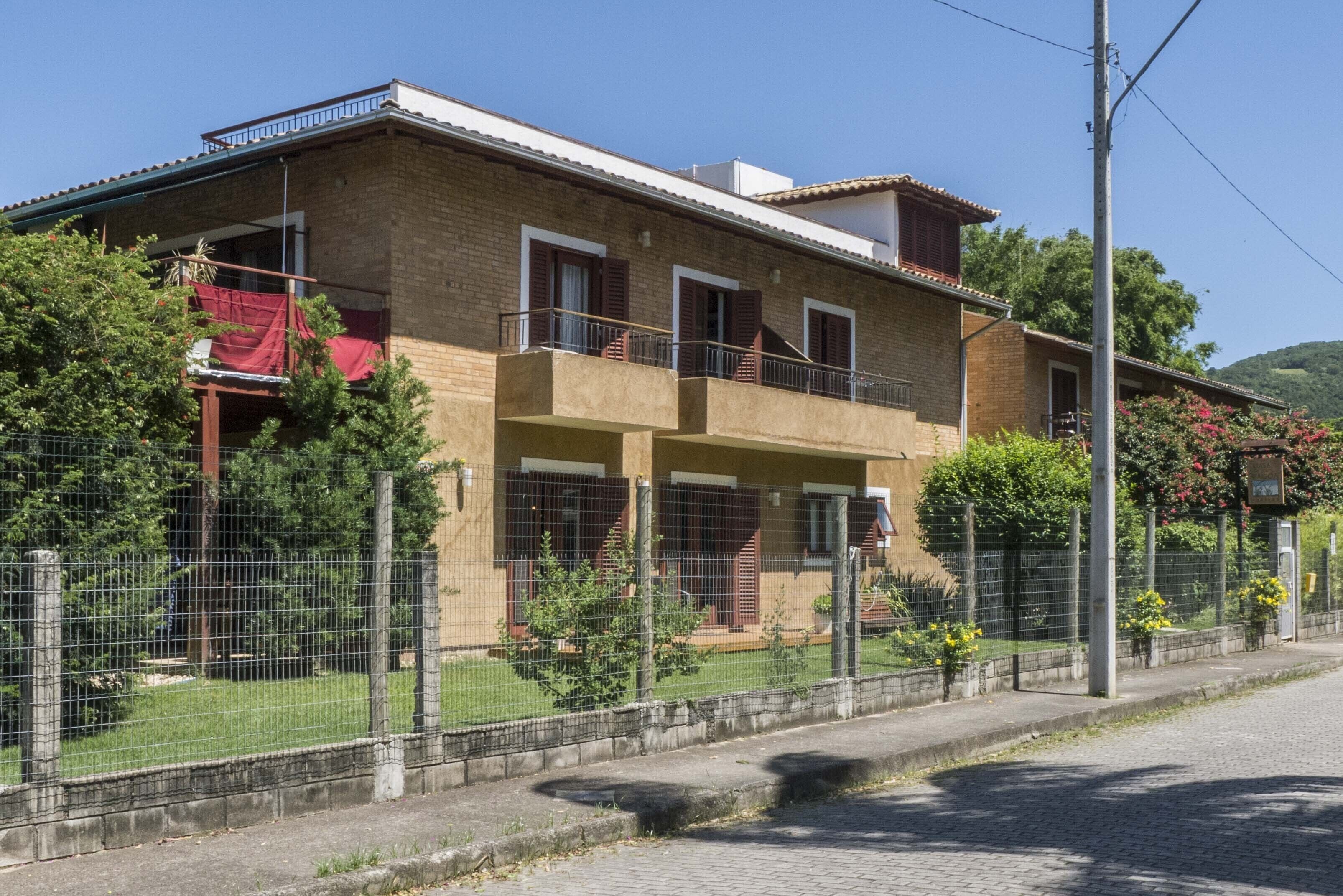 Edificio residencial de dos pisos con un balcón en el nivel superior, rodeado de una cerca y follaje.