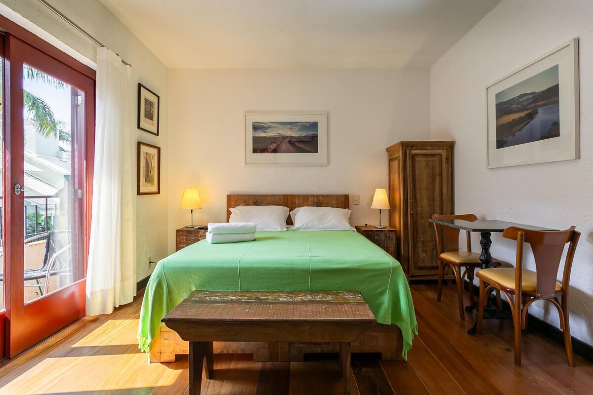 Ein Hotelzimmer mit Bett, einer Holzbank, einem Tisch mit Stühlen, einem Schrank aus Holz und Blick durch eine Glastür.