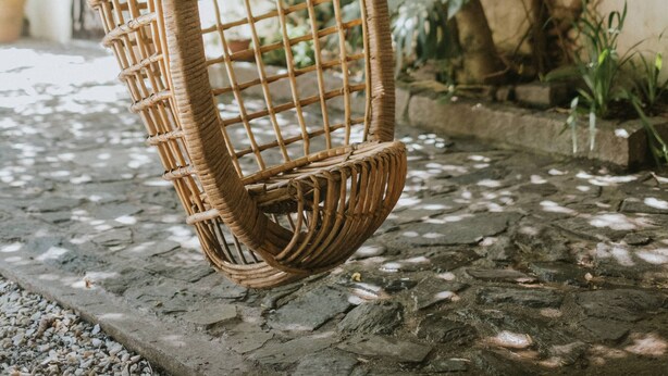 Una silla colgante de mimbre, suspendida de una cadena, ubicada en un jardín con suelo de piedra y abundante vegetación al fondo.