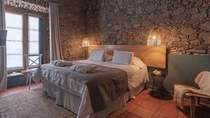 Un dormitorio con pared de piedra, una cama con ropa de cama blanca, dos lámparas de noche, una silla y una mesita con un teléfono.
