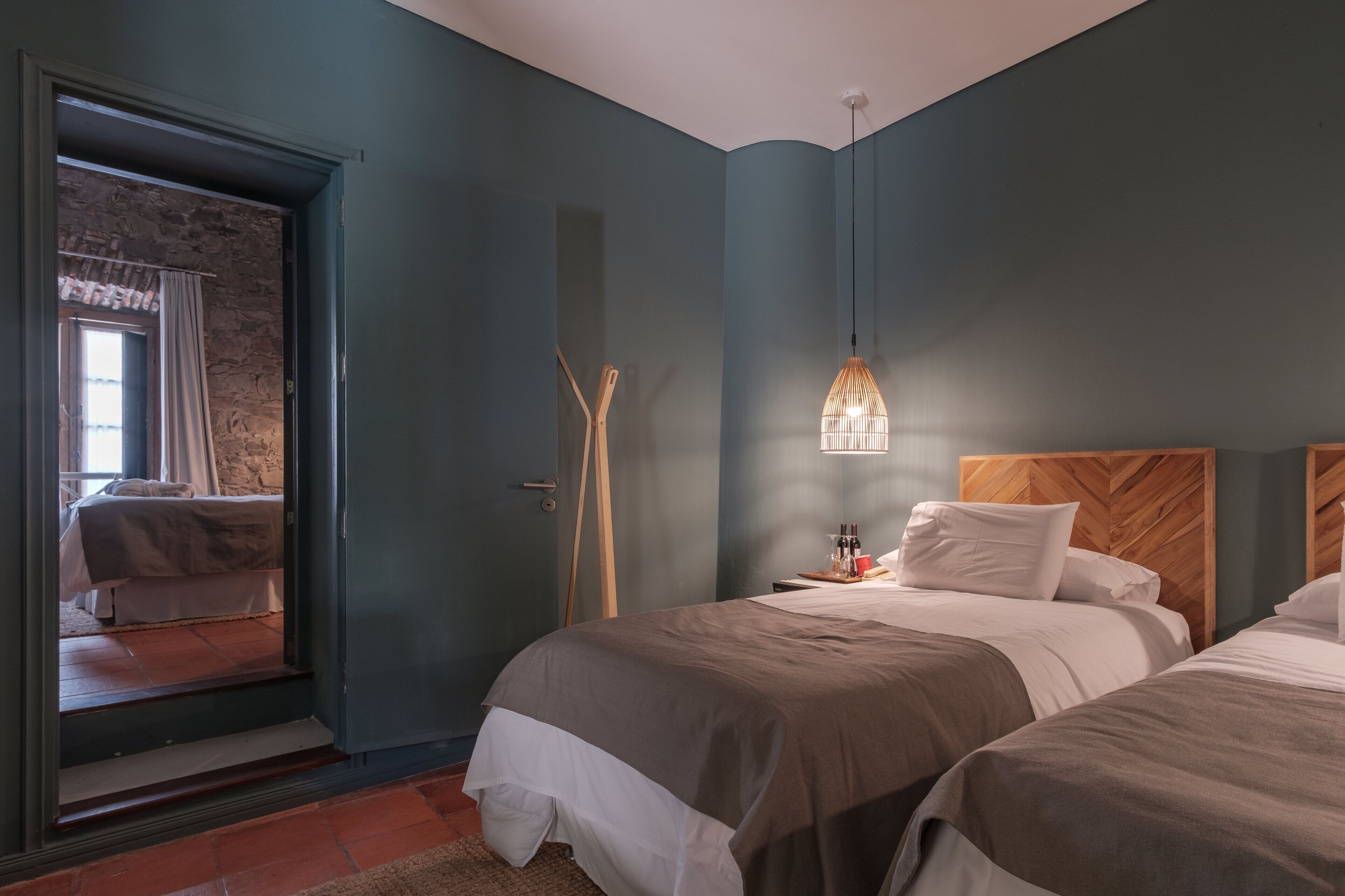 A bedroom with two beds, a wooden headboard, a hanging light fixture, and a view of a stone wall through an open door.