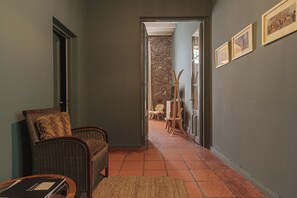 A hallway with a wicker chair, a wooden table, and a rug. There are framed pictures on the wall and a stone wall in the background.