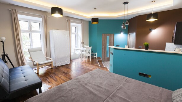 A modern hotel room with a bed, a seating area with a chair and table, a kitchenette, and a large window with curtains. - Fancy Ernesto II (Budapest)