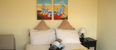 A hotel room with a bed, two pillows, a bedside table with a lamp, and a painting of a beach scene.