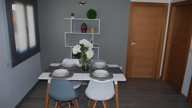 A dining area with a table set for four, two chairs, a vase with flowers, and a shelf with decorative items. - Apartamentos Rurales El Trillo (Malpartida de Plasencia)