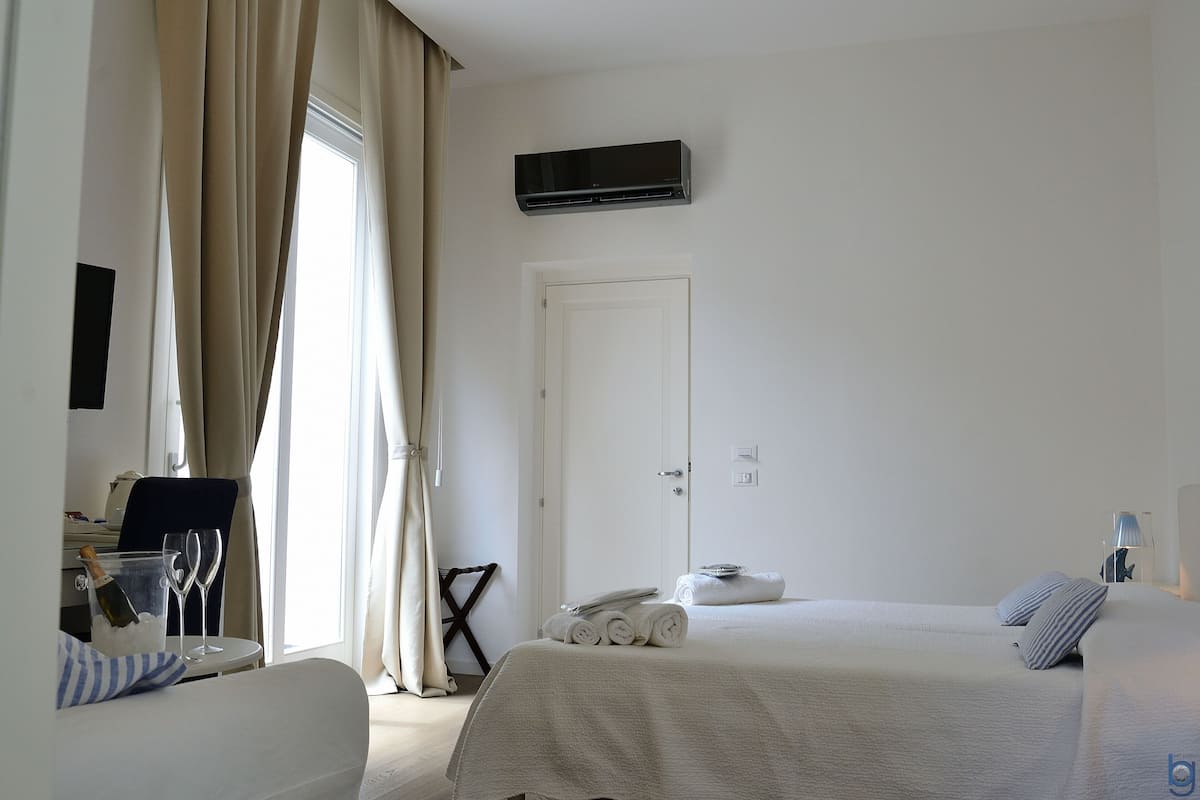 A hotel room with a bed, a chair, a table with a bottle and glasses, and a door.