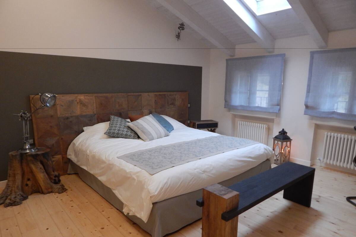 Een slaapkamer met een houten hoofdeinde, een bed met wit beddengoed, een bankje, een nachtkastje met een lamp en een raam met uitzicht.