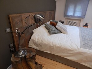 A bedroom with a wooden headboard, a bedside lamp, a bed with white bedding and patterned pillows, a wooden nightstand, and a radiator.