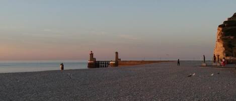A pebbled beach with a pier and lighthouses, a cliff, and people walking.
