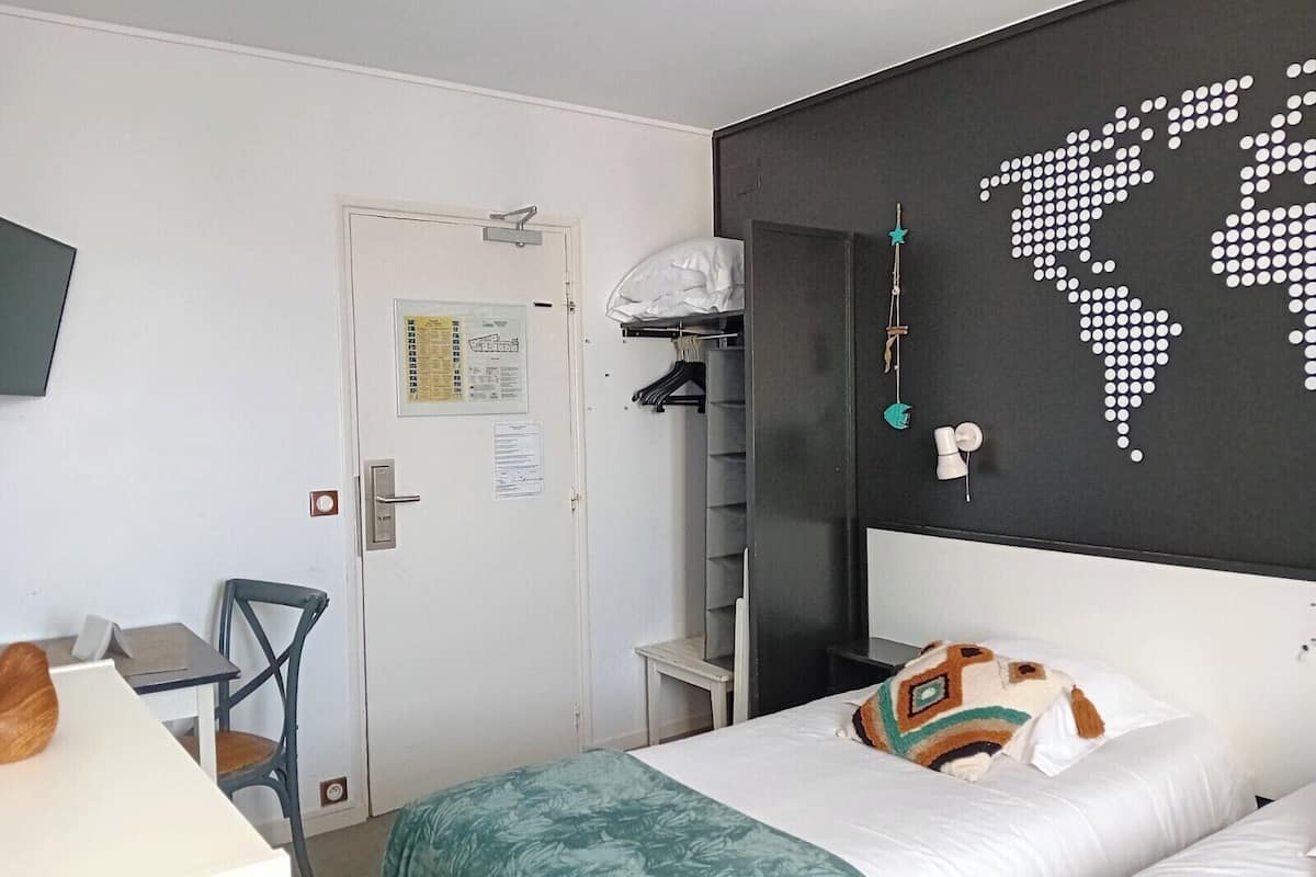 A hotel room with a bed, a desk, and a chair. There is a world map on the wall and a small shelf with a globe and a clock.