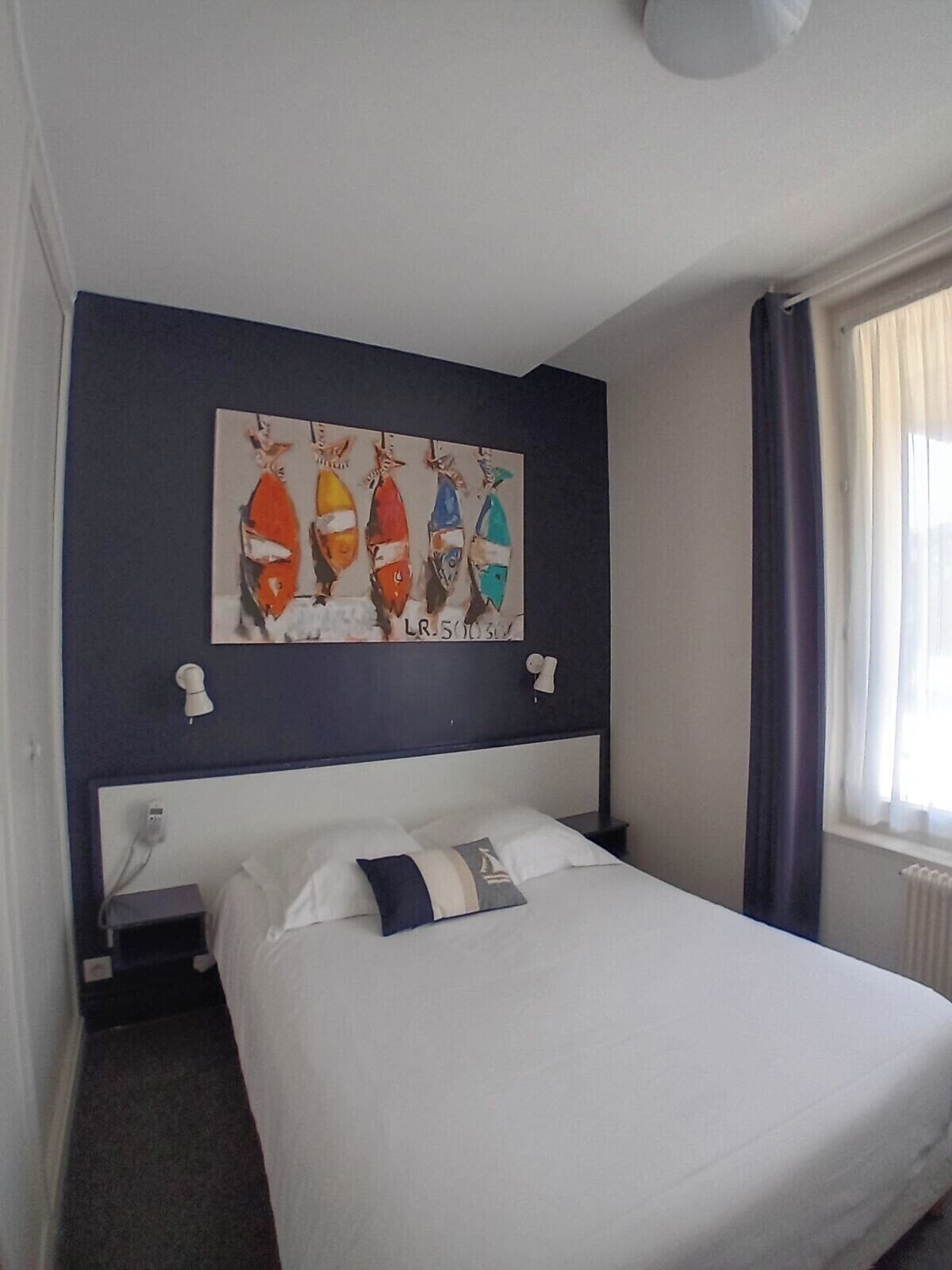 A hotel room with a bed, a nightstand, a wall-mounted painting of fish, and a window with curtains.