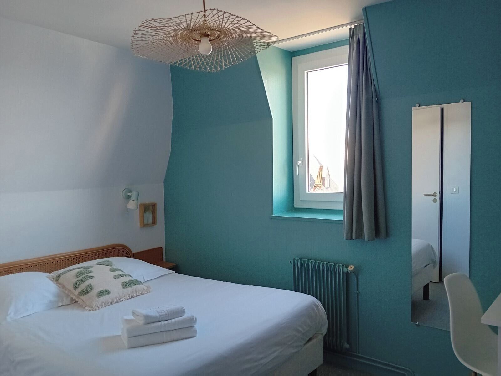Un dormitorio con cama, ventana con cortinas, radiador, espejo y lámpara colgante.