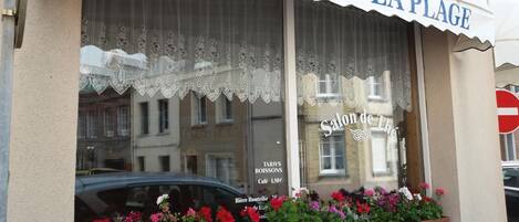 Fachada de hotel com placa indicando "HOTEL DE LA PLAGE" e um arranjo de flores em vasos na vitrine.