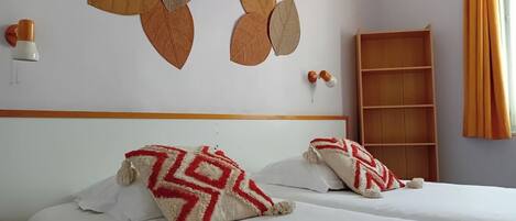 A hotel room with two beds, a wooden headboard, a wall-mounted shelf, and a window with curtains.