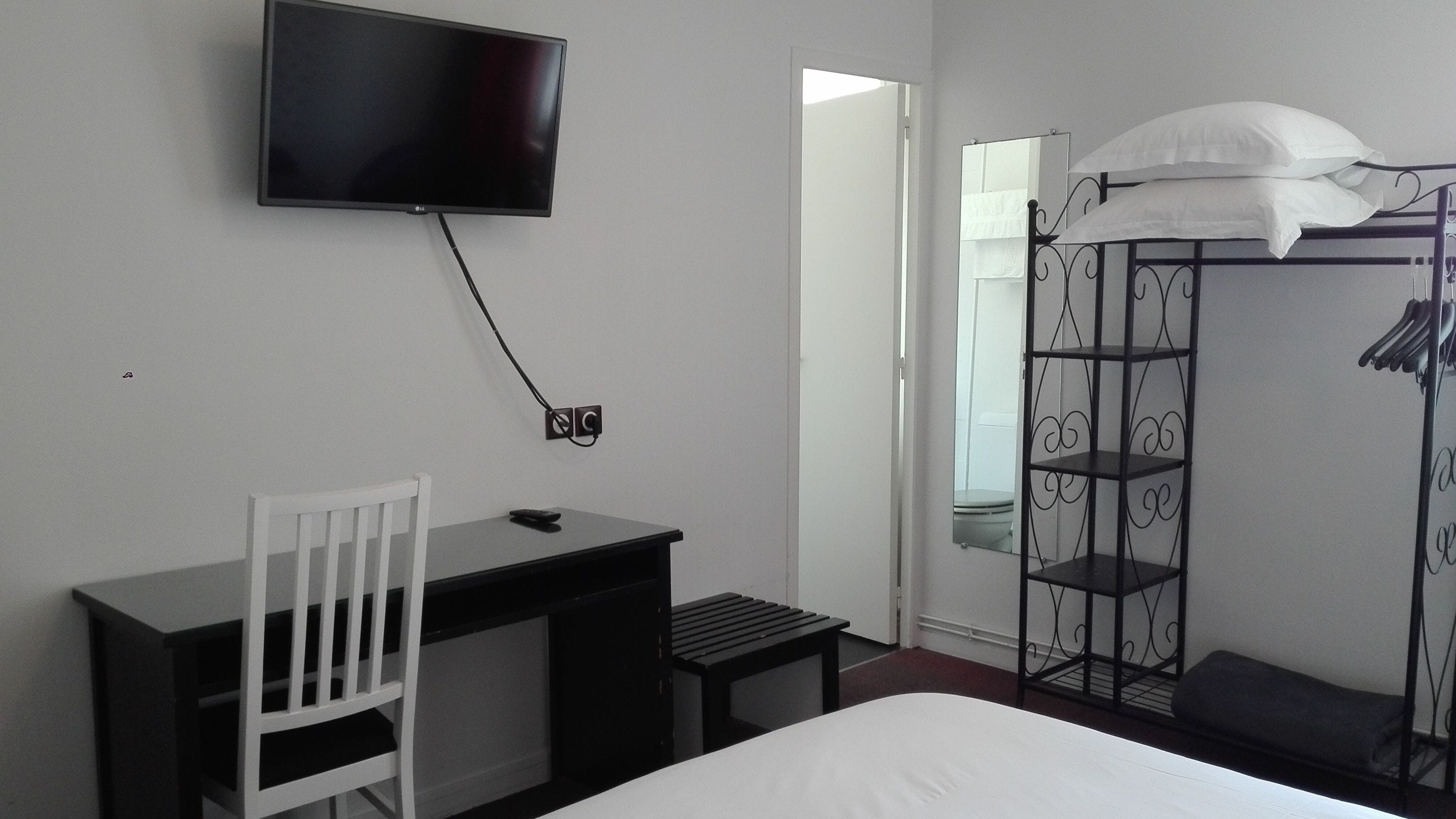 A hotel room with a bed, a desk, a chair, a TV mounted on the wall, a wardrobe with a mirror, and a bathroom visible through an open door.