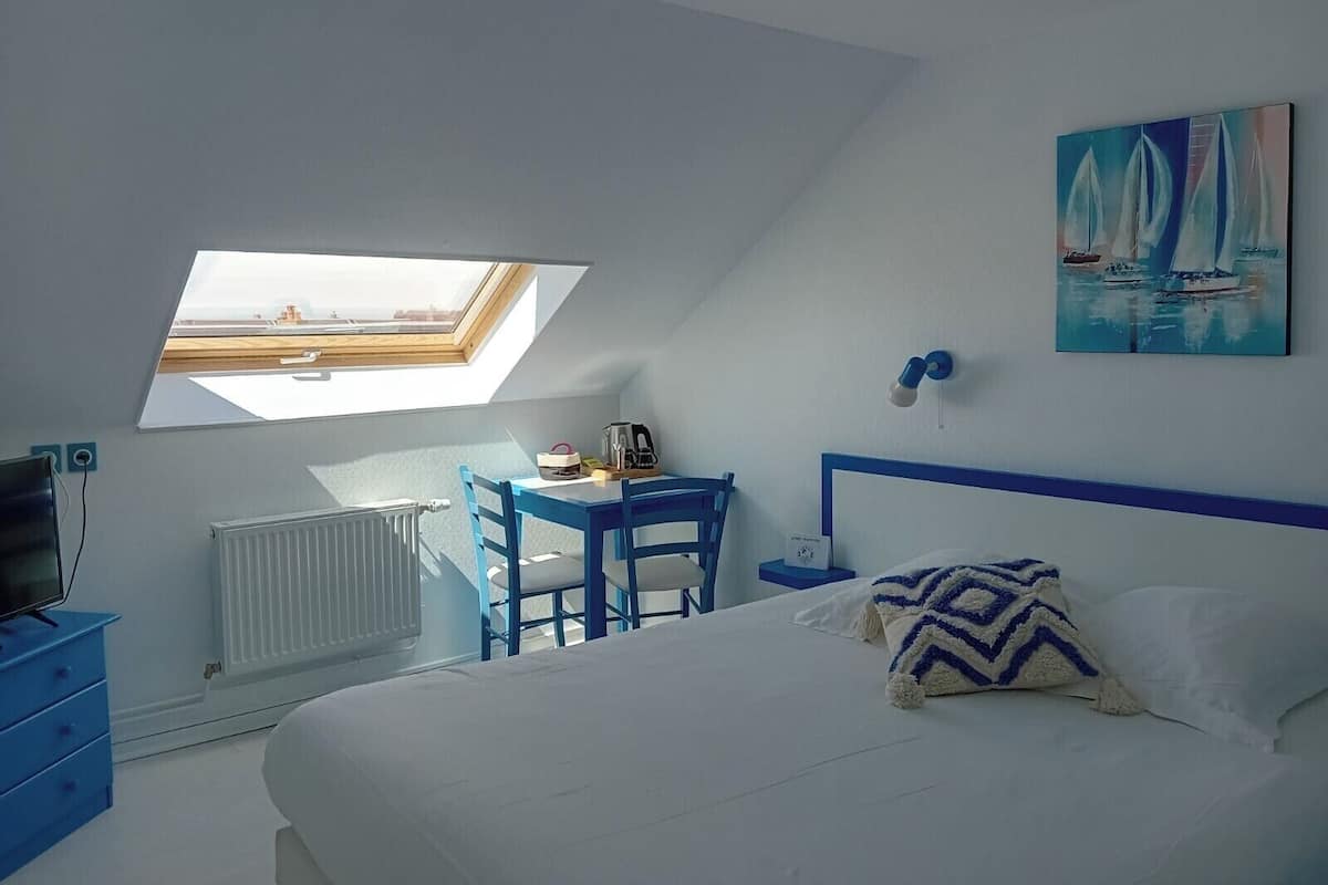 A bedroom with a skylight, a bed, a desk with a chair, a painting, and a nightstand.