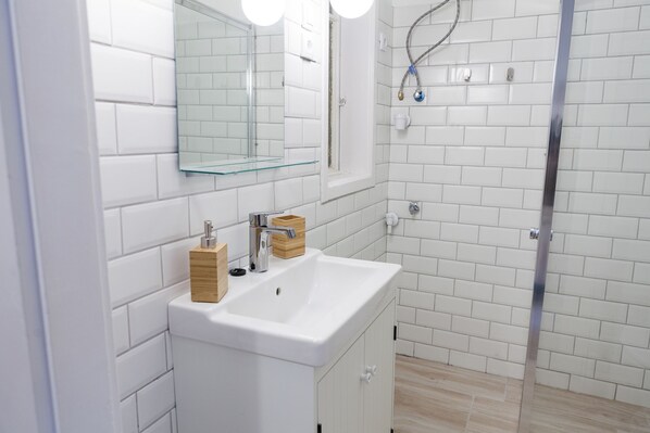 Un baño moderno con baldosas blancas tipo metro, mampara de vidrio, lavamanos blanco con dispensador de jabón de madera, y un espejo sobre el lavamanos.