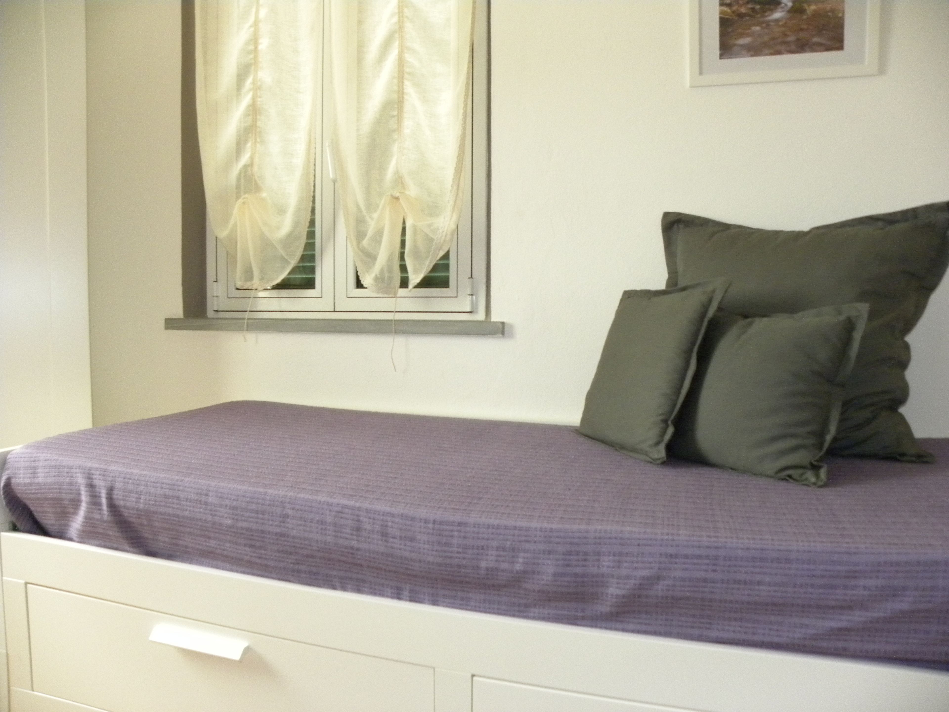 A bed with a purple bedspread, two green pillows, a window with white curtains, and a framed picture on the wall.