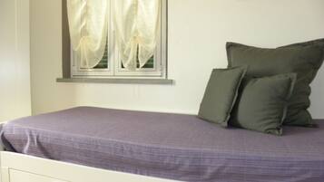 A bed with a purple bedspread, two green pillows, a window with white curtains, and a framed picture on the wall.