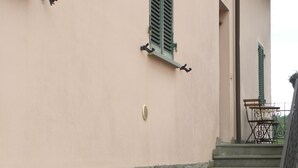 Ein Gebäude mit rustikalem Sockel, einer rosafarbenen oberen Wand und einer Treppe, die zu einem Balkon mit Gartenmöbeln führt.