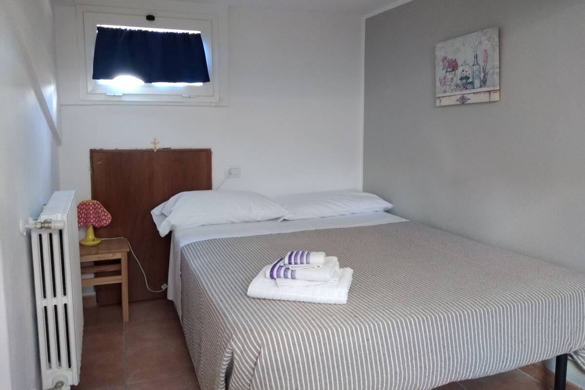 Habitación individual con ventana, radiador, una mesita con lámpara y un cuadro en la pared.