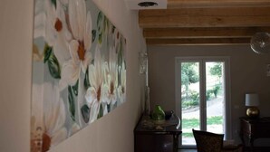 Una habitación con un cuadro grande de flores en la pared, techo de madera, ventana con vistas a la vegetación y una mesa con una lámpara.