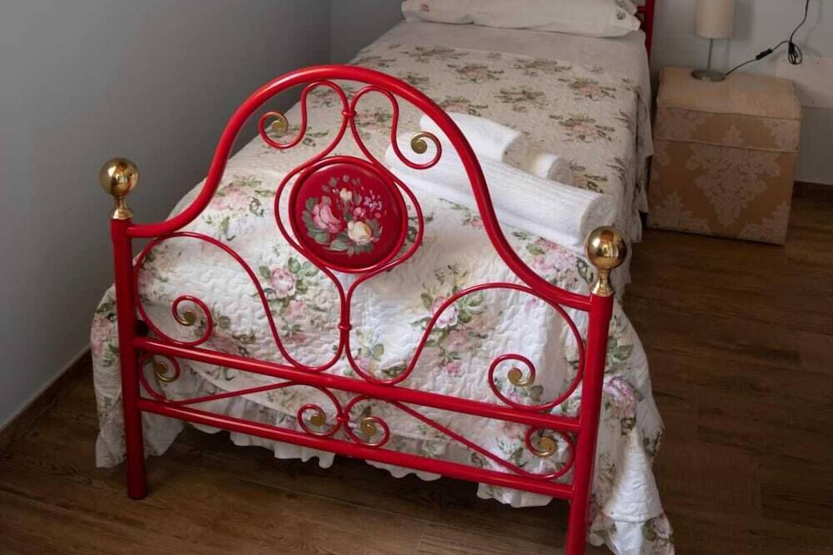 Ein kleines Zimmer mit einem Einzelbett, einem roten Metallgestell, floralem Bettbezug, einem Spiegel an der Wand und einem Nachttisch mit Lampe.