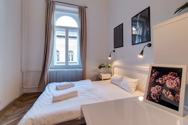 A neatly made bed with white linens and a towel, a bedside table with a lamp, a framed picture of flowers, and a window with curtains.