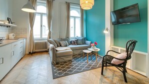 A modern living room with a sofa, a small table, and a chair. There is a TV mounted on the wall and a window with curtains. - Highlight Ernesto II (Budapest)