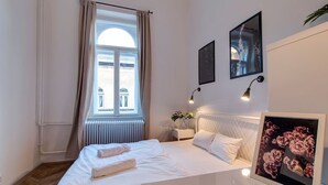 Una cama bien tendida con sábanas blancas y una toalla, una mesita de noche con una lámpara, un cuadro con flores y una ventana con cortinas.