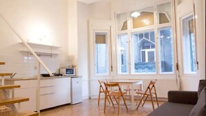 A modern kitchen with white cabinets and a microwave, a dining area with wooden chairs, and a large window with a view of a building outside. - Incredible Ernesto I (Budapest)