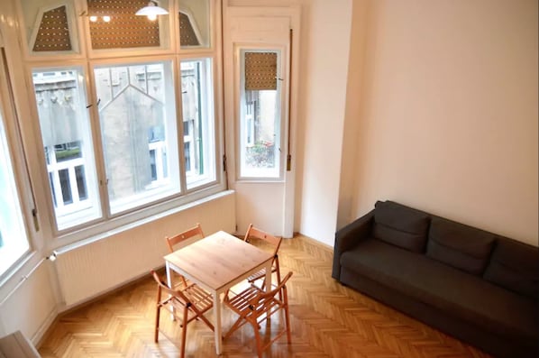 A room with a wooden herringbone floor, a dining table with chairs, a dark-colored sofa, and large windows with blinds.