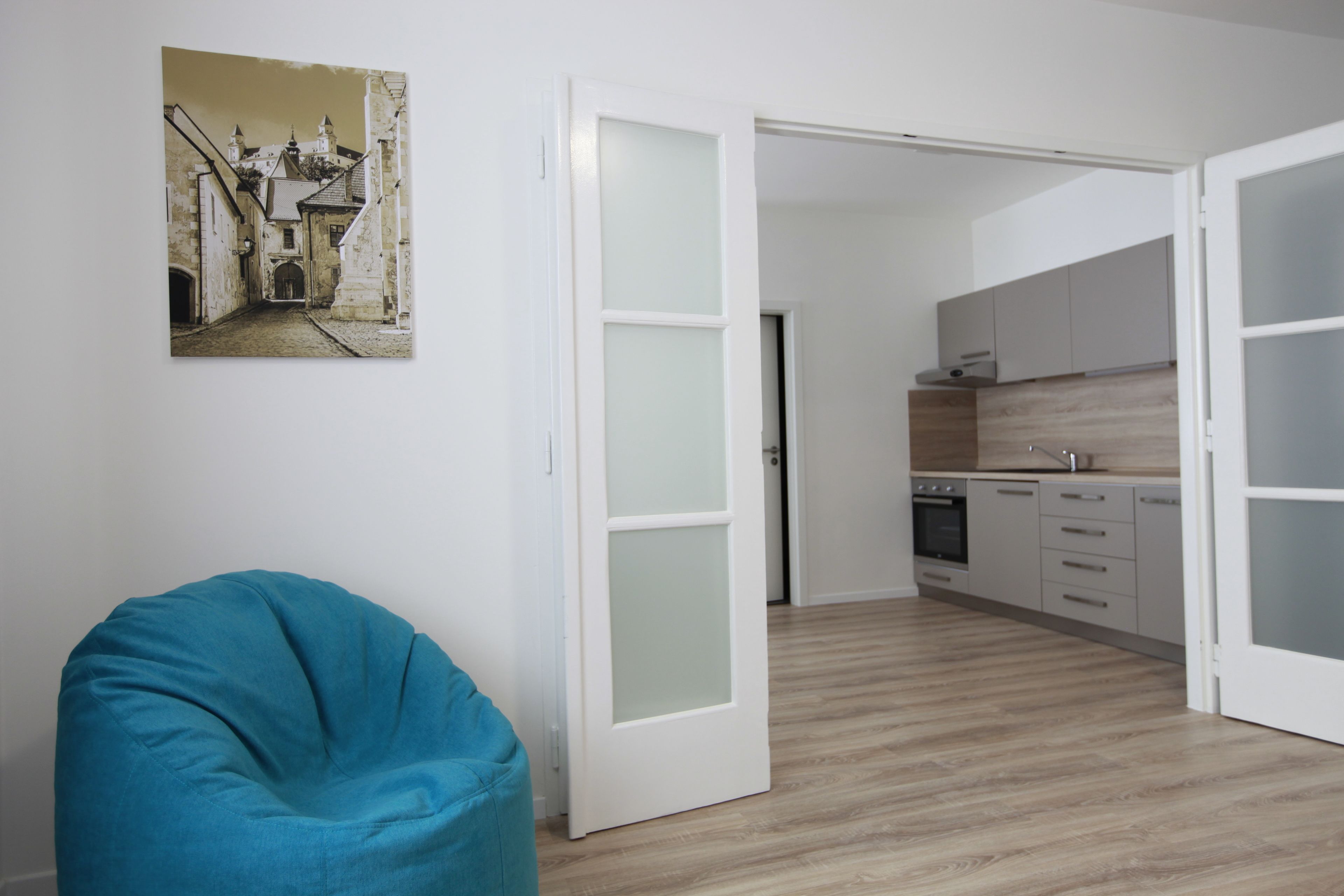Een moderne keuken met witte kastjes en een houten vloer, een blauwgroene kruurzak en een lijst met een foto van een straatbeeld.