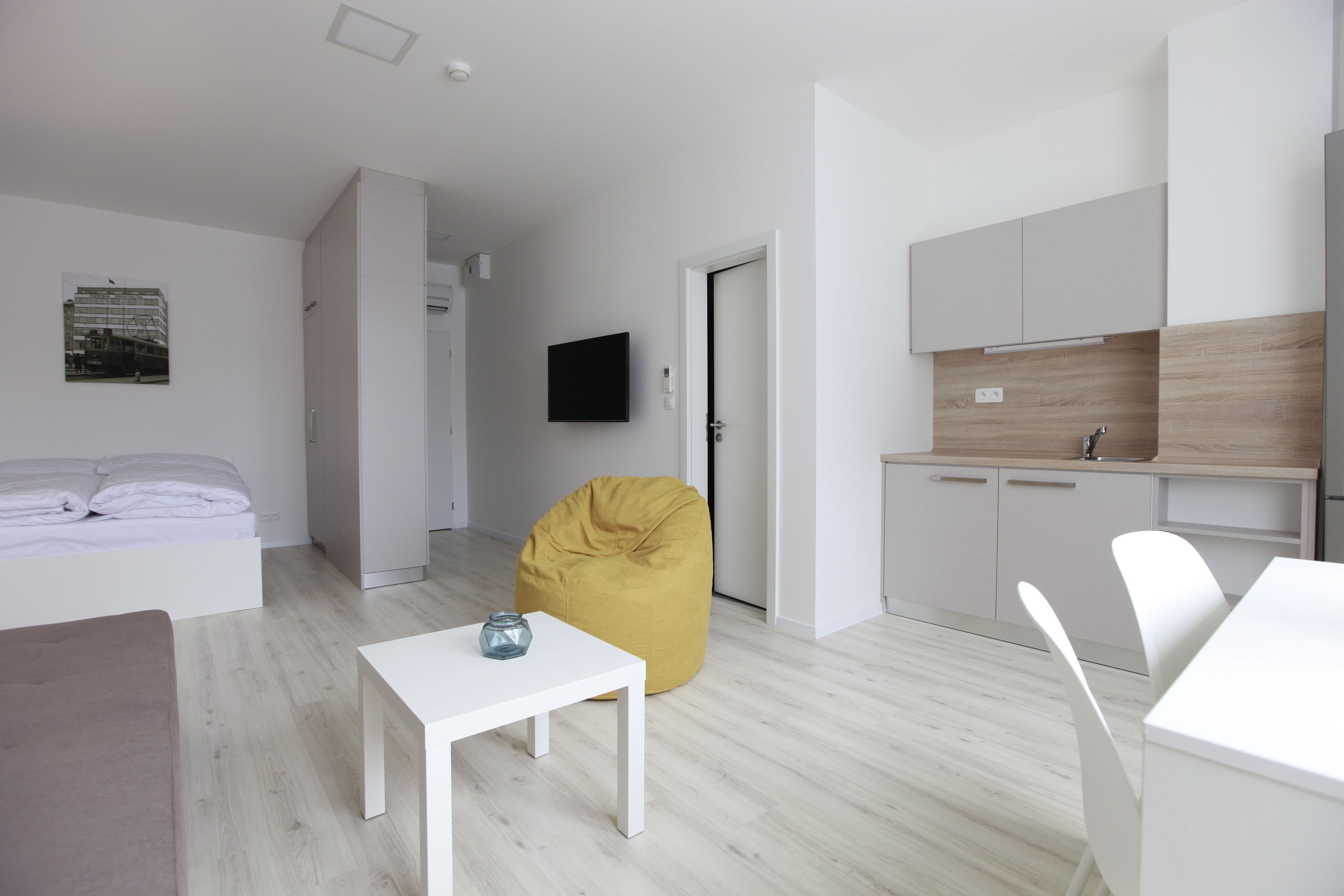 A modern, minimalist living room with a sofa, a white coffee table, and a yellow bean bag. There is a kitchen area with white cabinets and a flat-screen TV mounted on the wall.