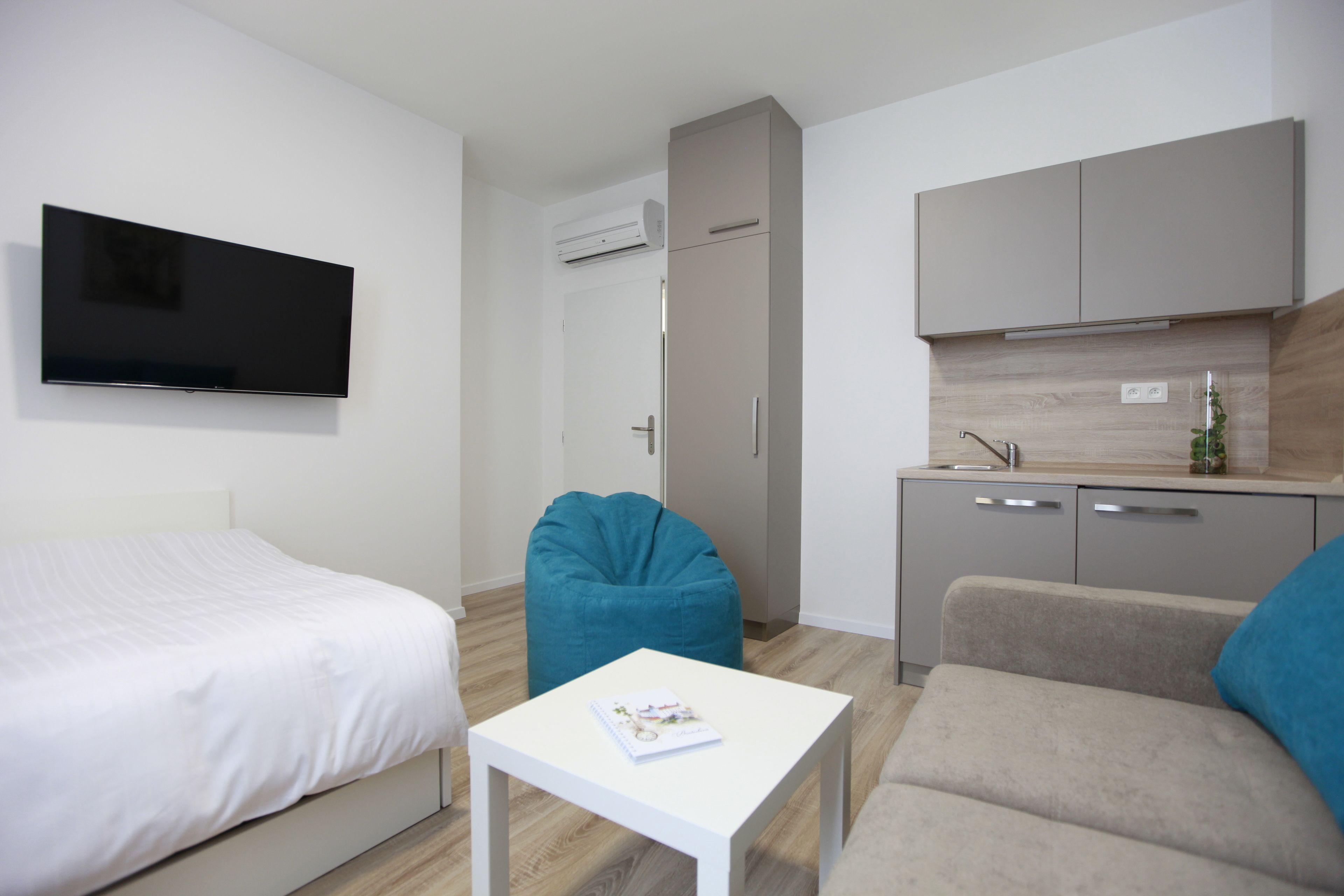 Camera d'albergo moderna con un letto, un piccolo tavolo bianco, una poltrona a sacco color petrolio, un angolo cottura con lavello e una TV a schermo piatto fissata al muro.