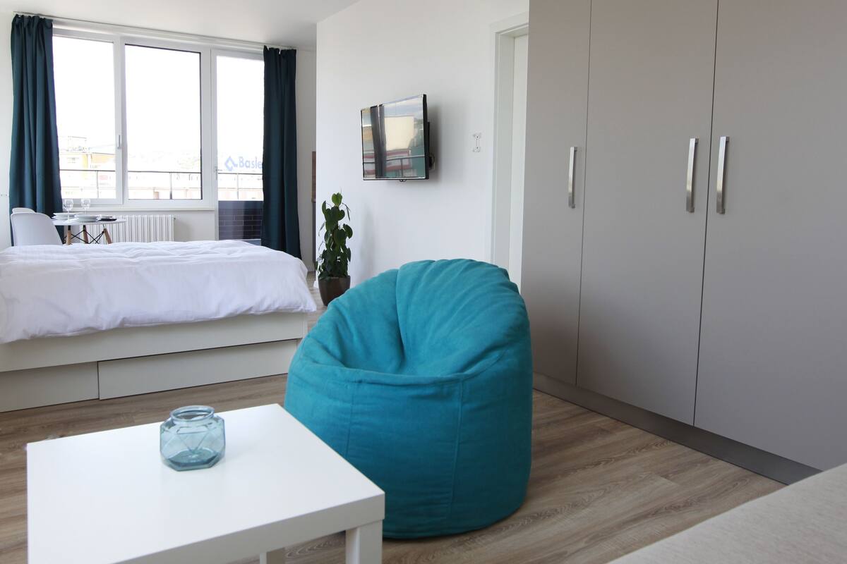 Ein modernes Schlafzimmer mit einem Bett, einem türkisfarbenen Sitzsack, einem weißen Tisch mit einem Glasbehälter und einem Fernseher an der Wand.