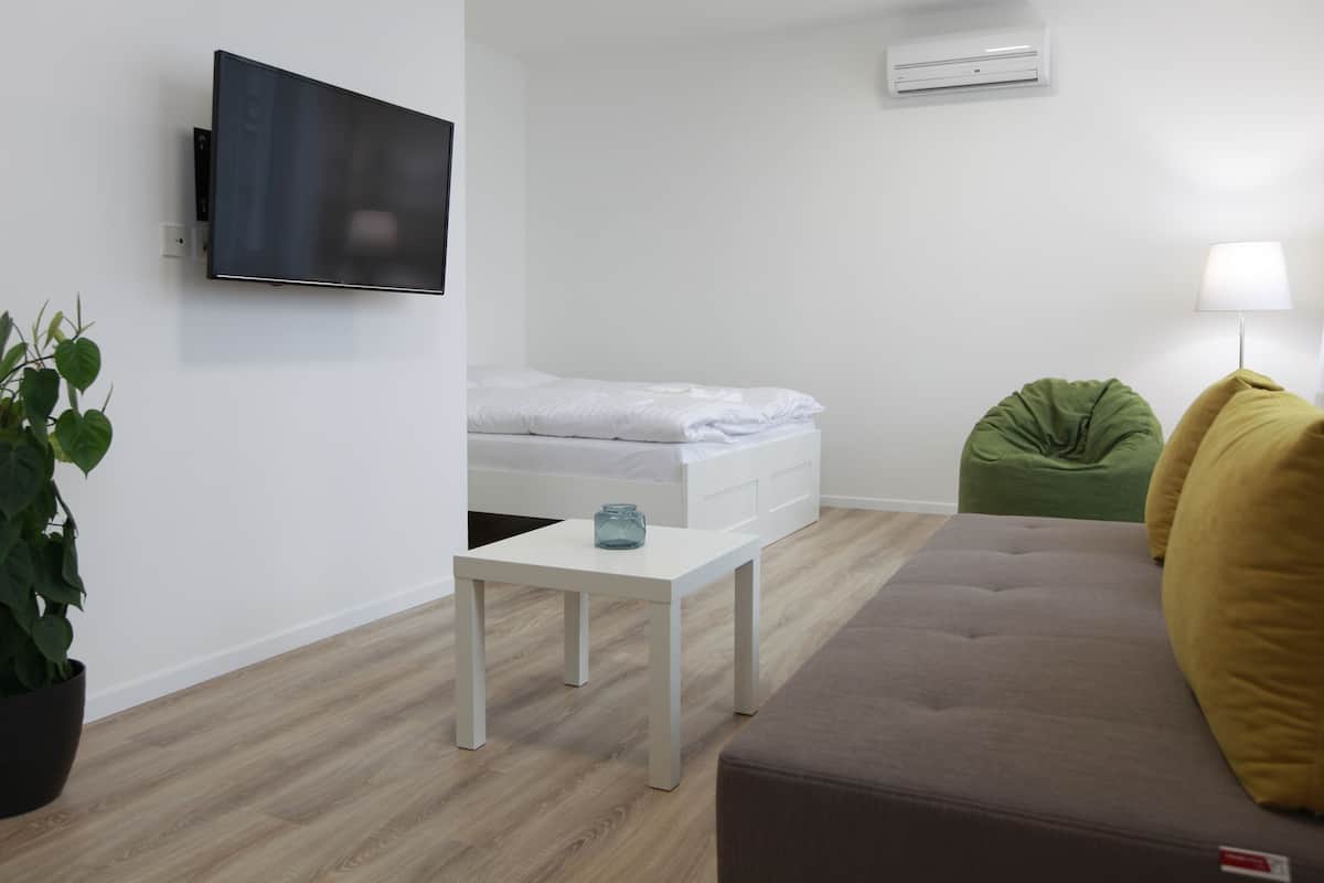 Ein modernes, minimalistisches Zimmer mit einem an der Wand montierten Flachbildfernseher, einem Bett mit weißer Bettwäsche, einem kleinen weißen Tisch, einem grünen Sitzsack, einem braunen Sofa und einer Stehlampe.