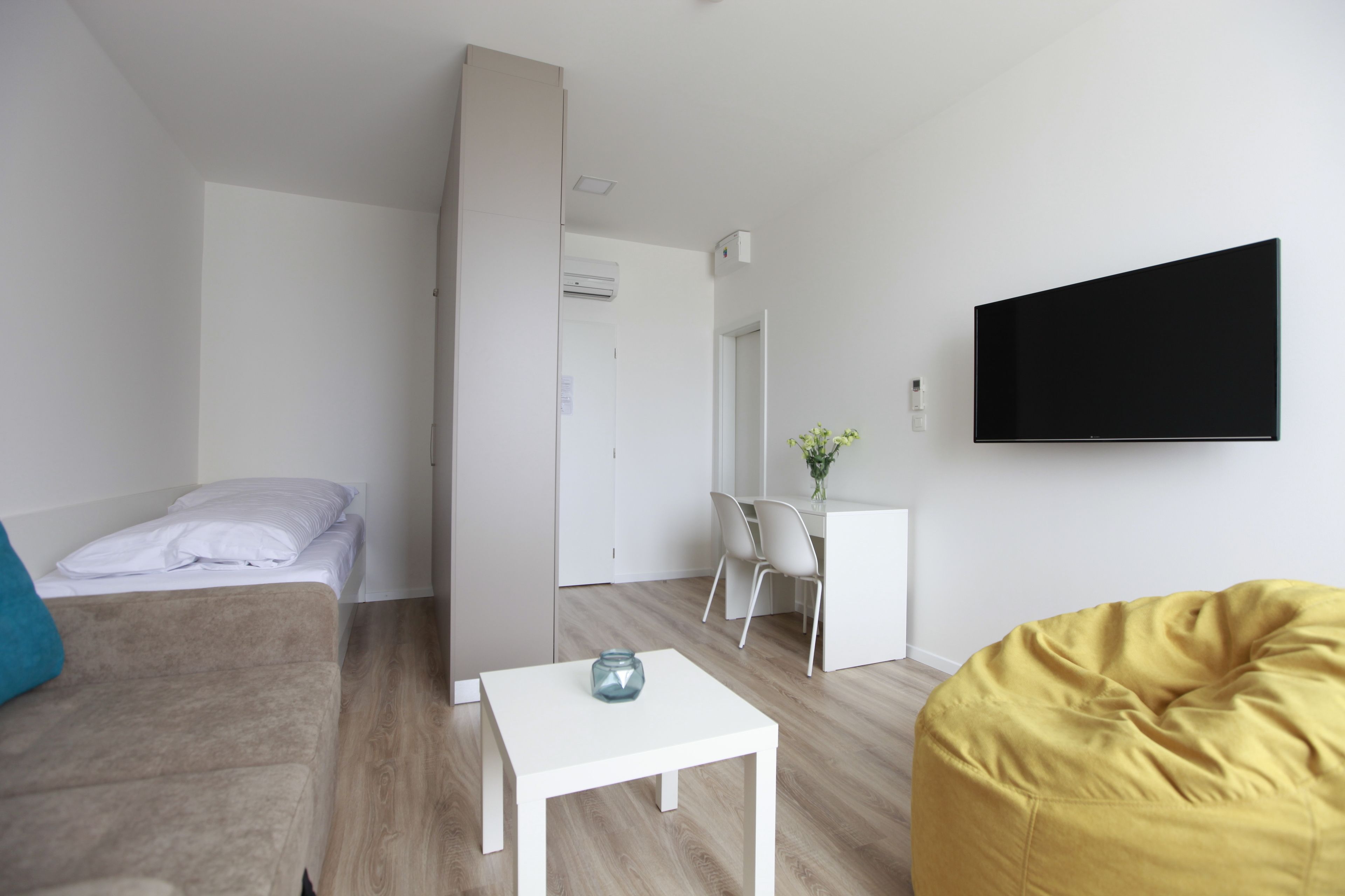 A modern living room with a sofa, a yellow bean bag, a white coffee table, and a flat-screen TV mounted on the wall.