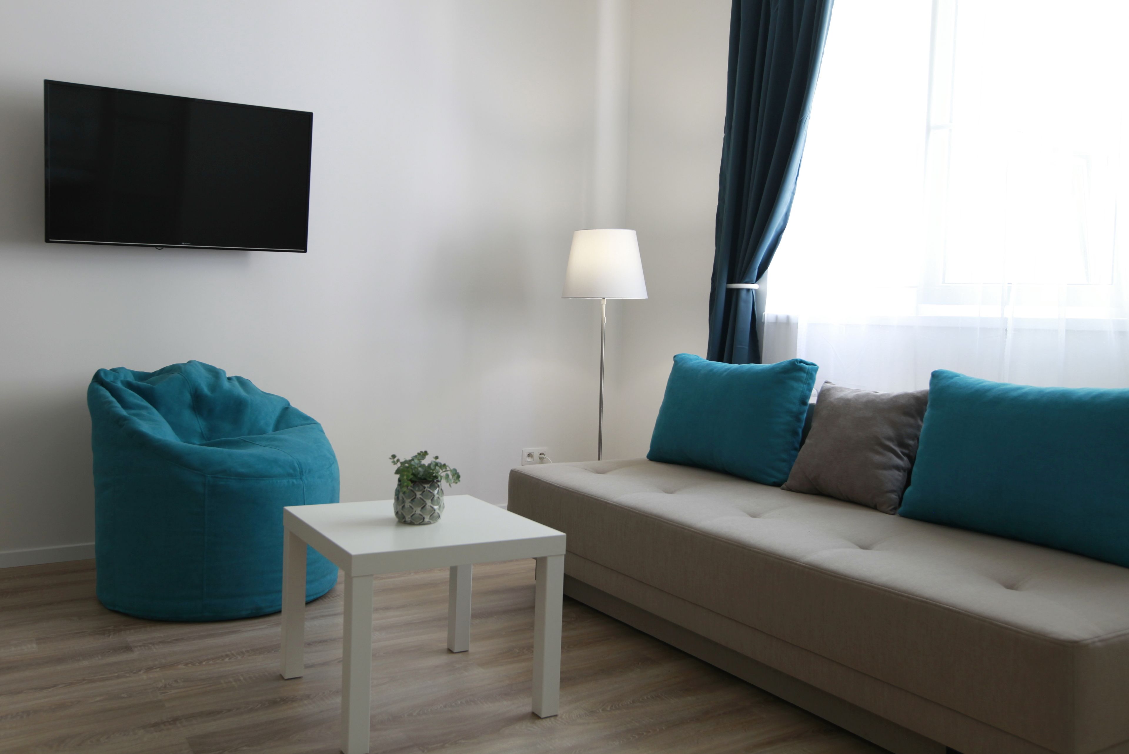 Una sala de estar moderna con un televisor de pantalla plana, un sillón tipo cojín color turquesa, una mesa de centro blanca con una planta, un sofá color beige con cojines turquesa y marrón, y una lámpara de pie.