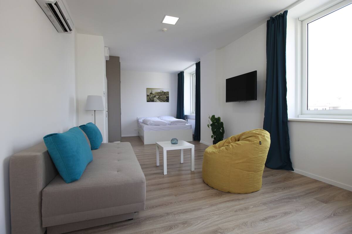 Ein modernes, helles Zimmer mit einer Couch, einem kleinen Tisch, einem Sitzsack, einem Bett und einem Fernseher.