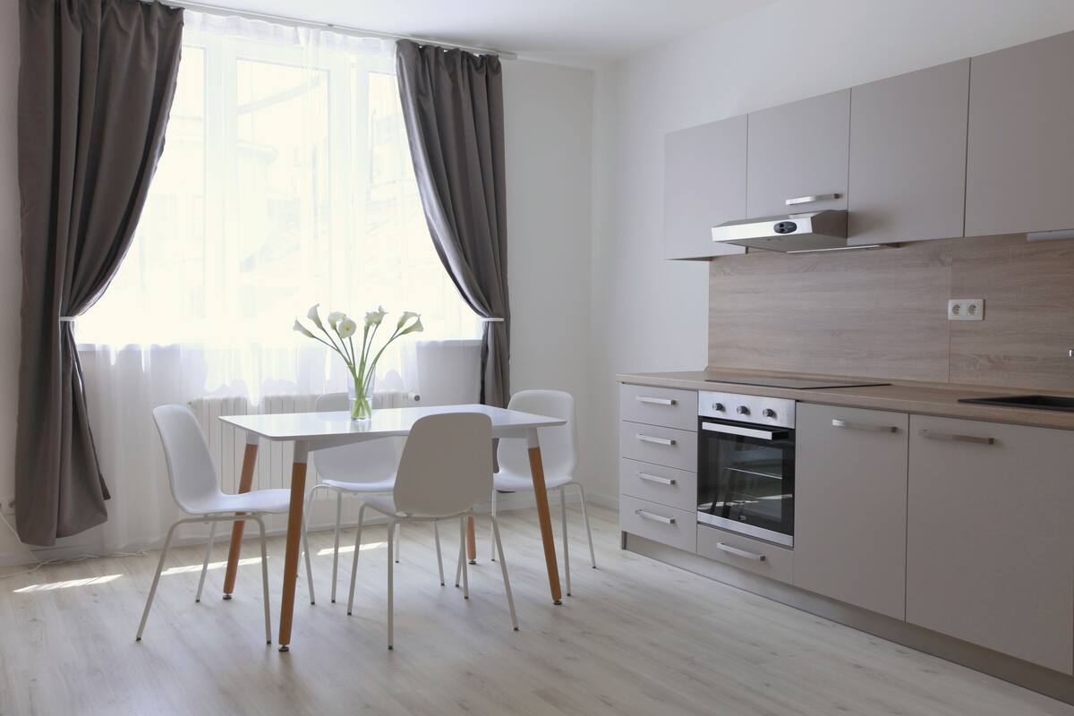 Eine moderne Küche mit Essbereich, ausgestattet mit einem weißen Tisch, Stühlen und einem großen Fenster mit Vorhängen.